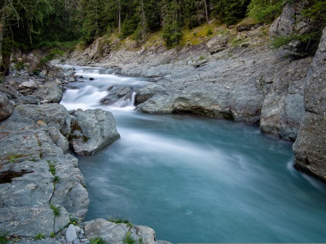 A mountain stream in long exposure: white rapids break over rocks above, then the water settles into a smooth, even turquoise flow below.