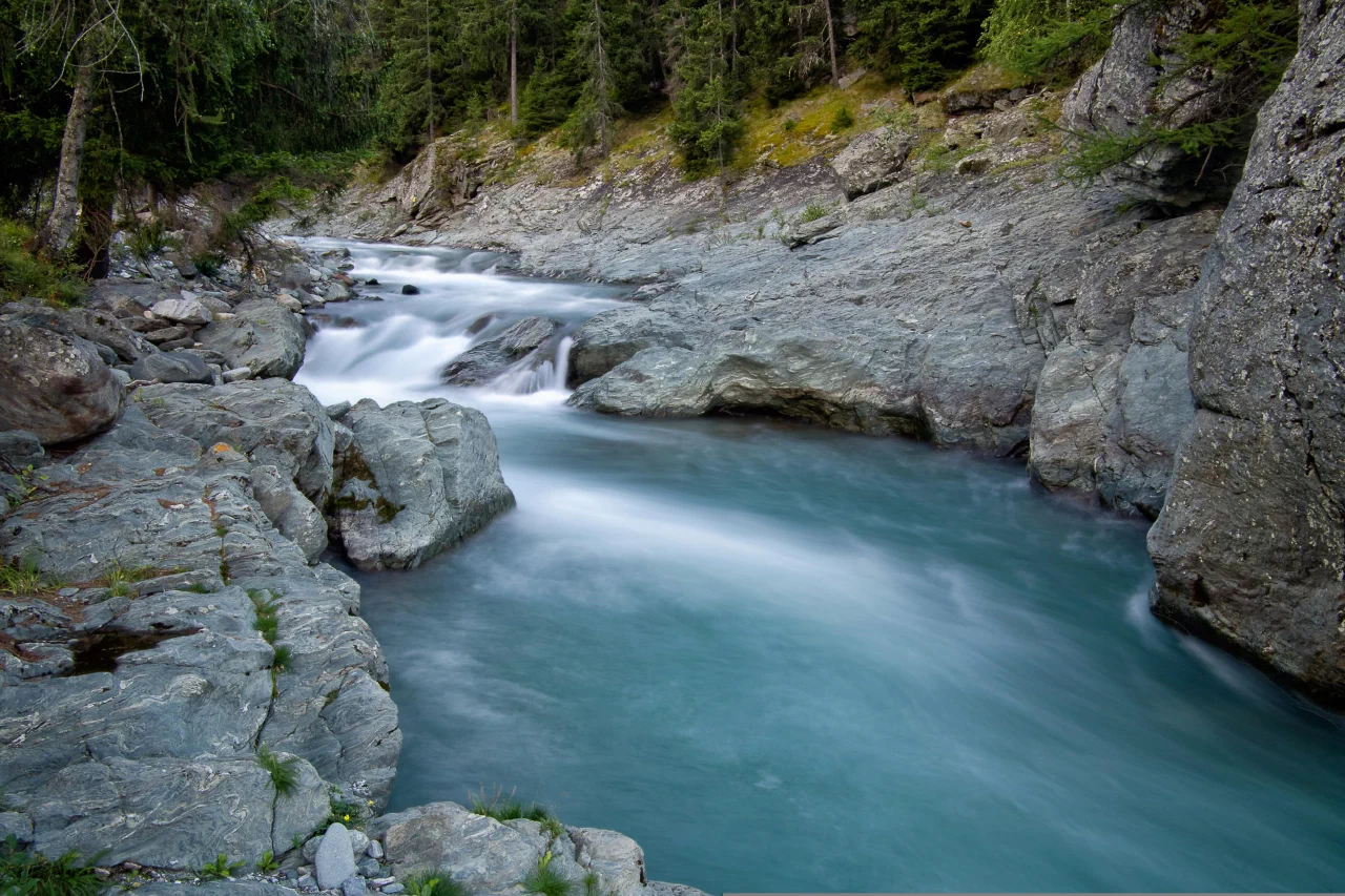 A mountain stream in long exposure: white rapids break over rocks above, then the water settles into a smooth, even turquoise flow below.